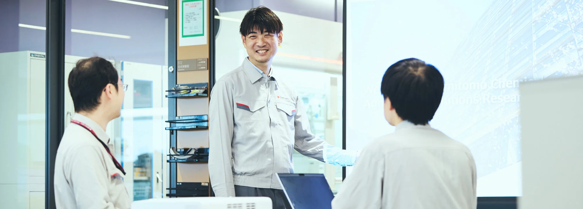 研究室で同僚に説明するK. M.